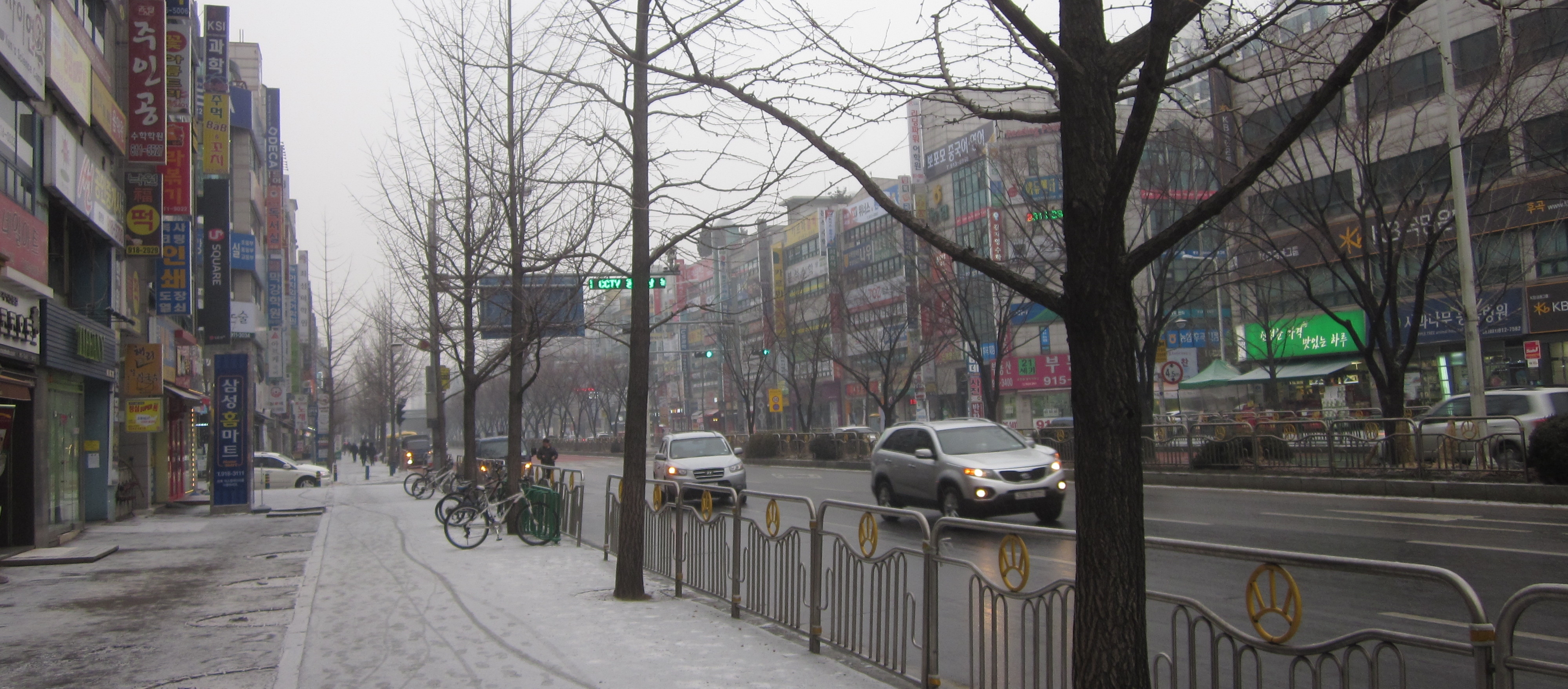 A picture taken in 2012 along a city street called Ilsan-ro in Goyang City, South Korea. Snow, some urban trees without leaves, lots of buildings along each side.