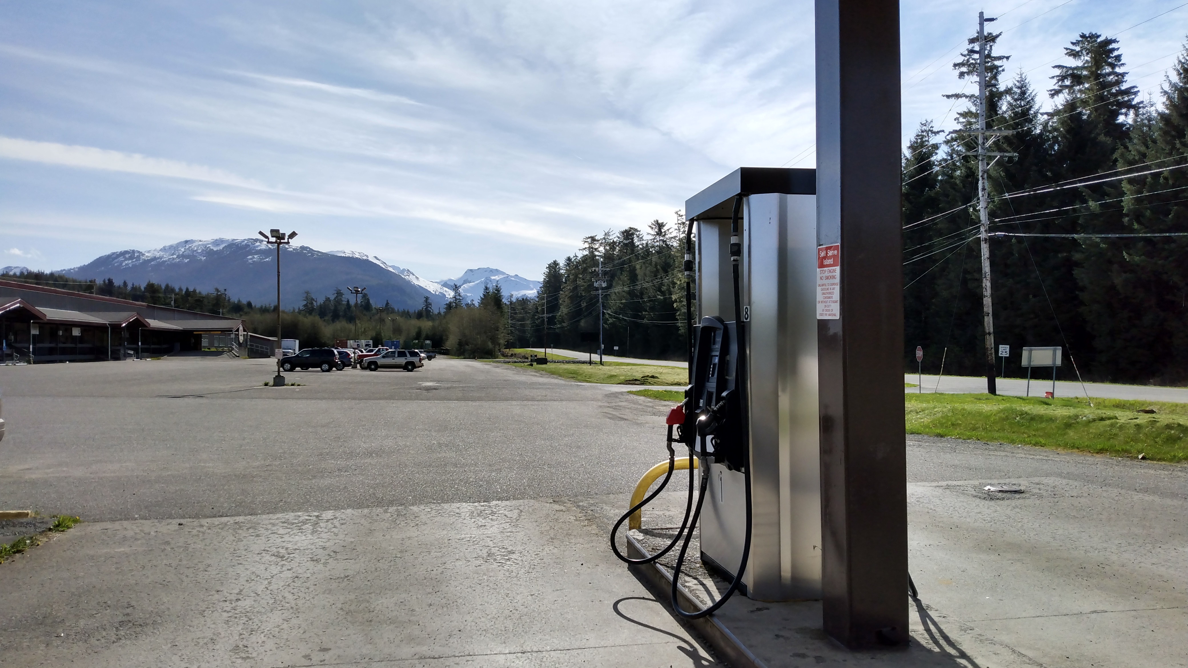 A gas-station pump in the foreground, with semi-desolate a parking lot in the middle ground and highway and trees and snowy mountains in the receding farther distance