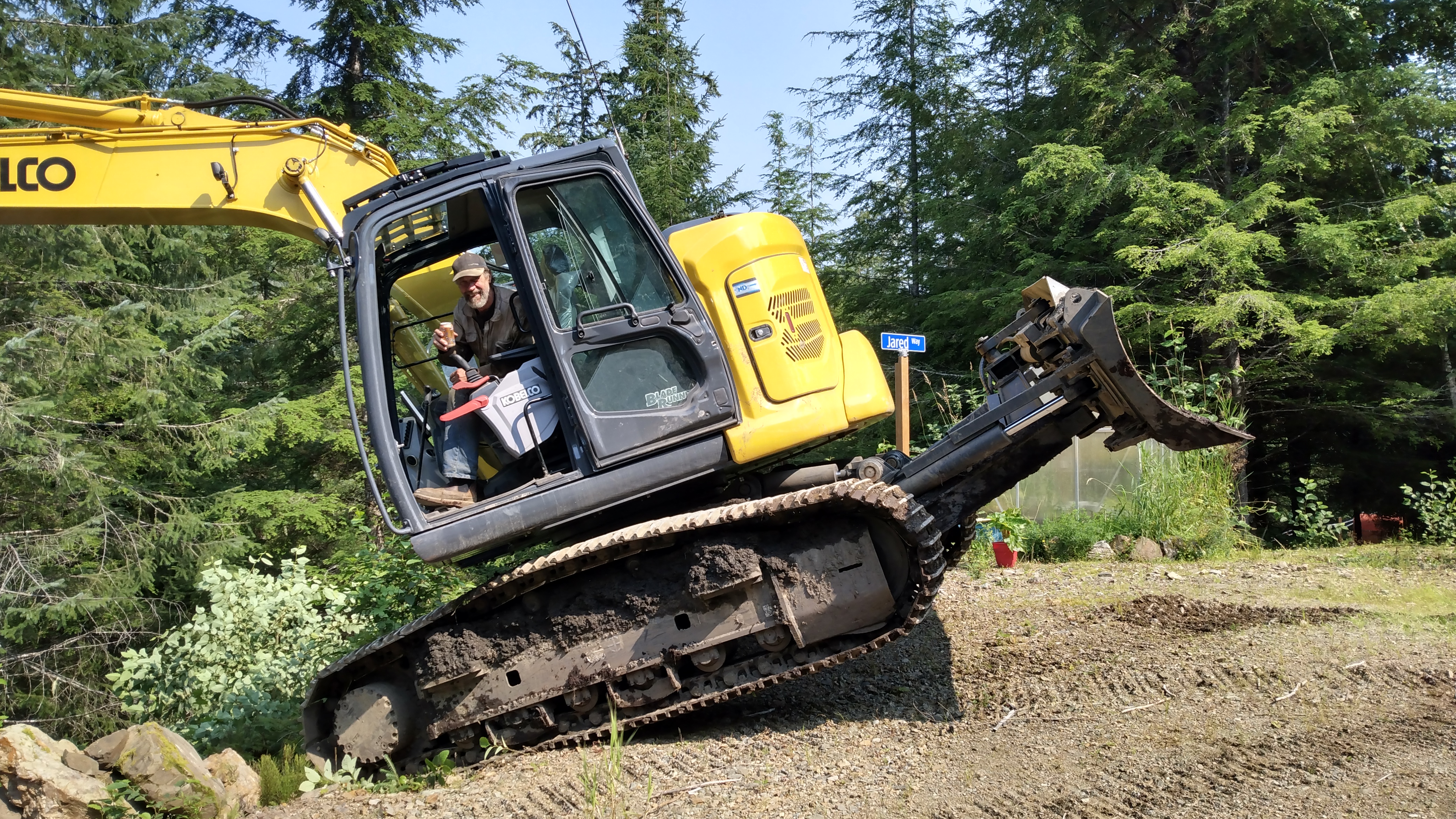 A closer view of the excavator with a grinning operator inside, holding a can of soda, and the excavator is balanced on the front of its tracks as it appears about to descend a steep hillside
