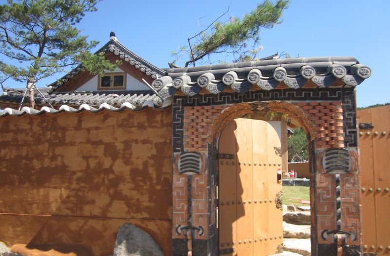 A wall with a gate of a traditional Korean style, in front of house and tree seen partially over the top of the wall